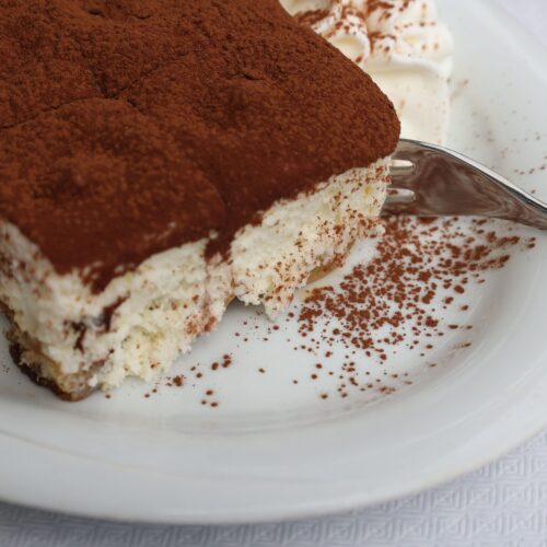 An appetizing slice of tiramisu covered in cocoa powder on a white plate.
